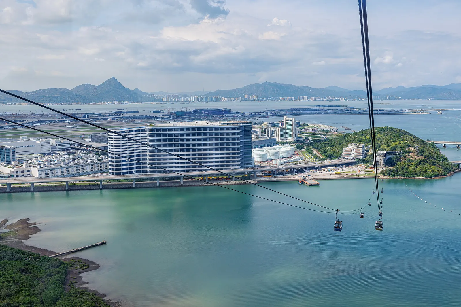 Looking down from a cabin over the green water of Tung Chung Bay to a white airport-side hotel, a wooded headland and the airport beyond, with cabins on the cable below