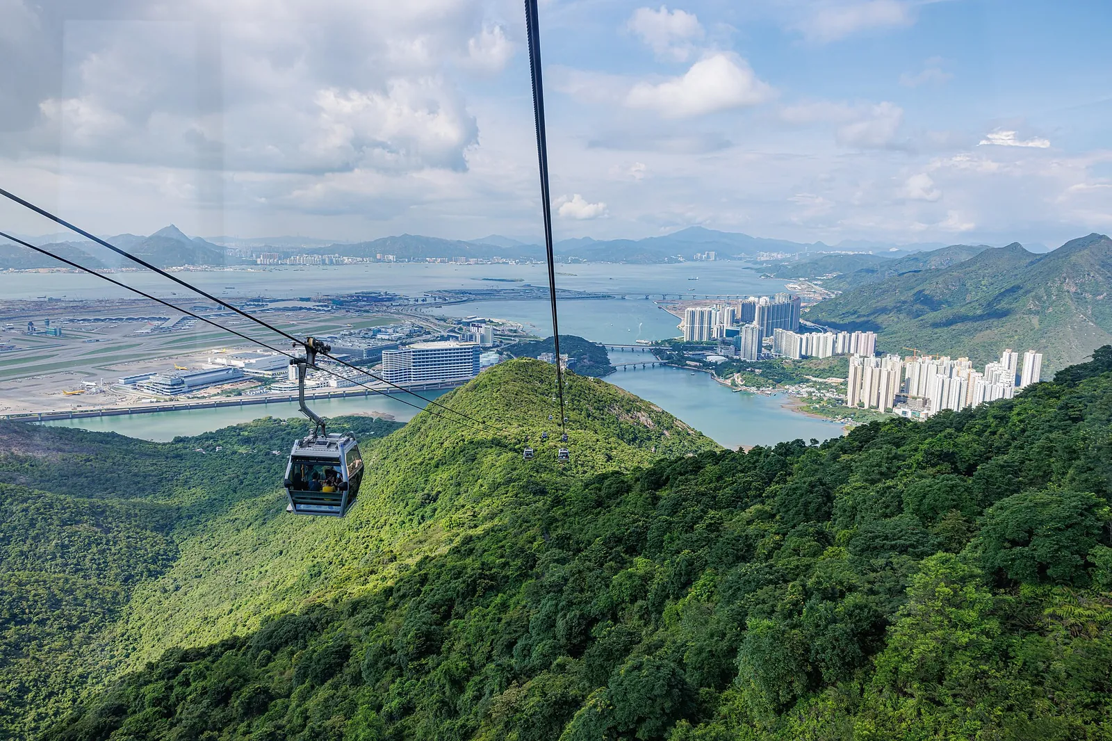 A grey cabin hangs from the cable above forested ridges, with Tung Chung's apartment towers, the bay and the airport runways stretching away below