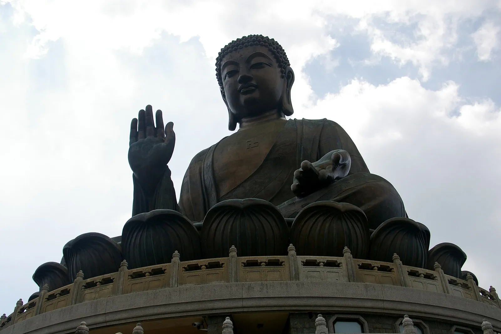 The bronze Tian Tan Buddha seen from below, one hand raised, seated on a lotus throne against a cloudy sky