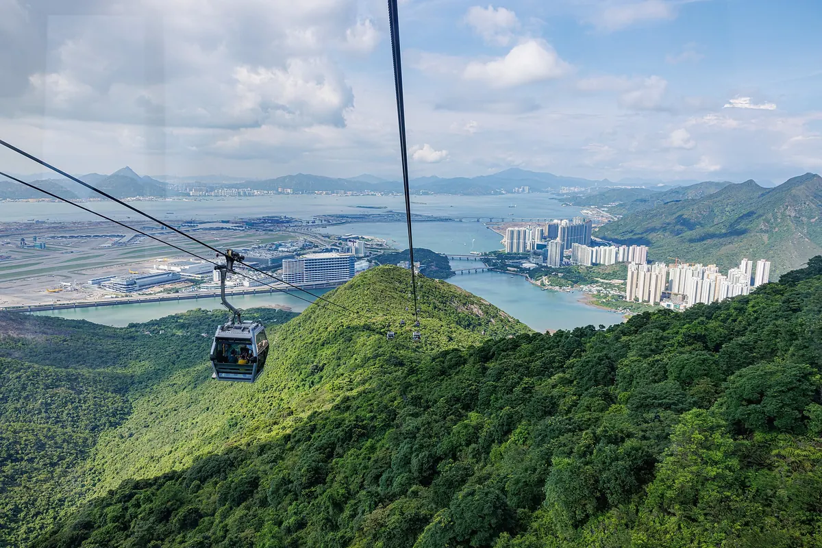 A grey cabin hangs from the cable above forested ridges, with Tung Chung's apartment towers, the bay and the airport runways stretching away below