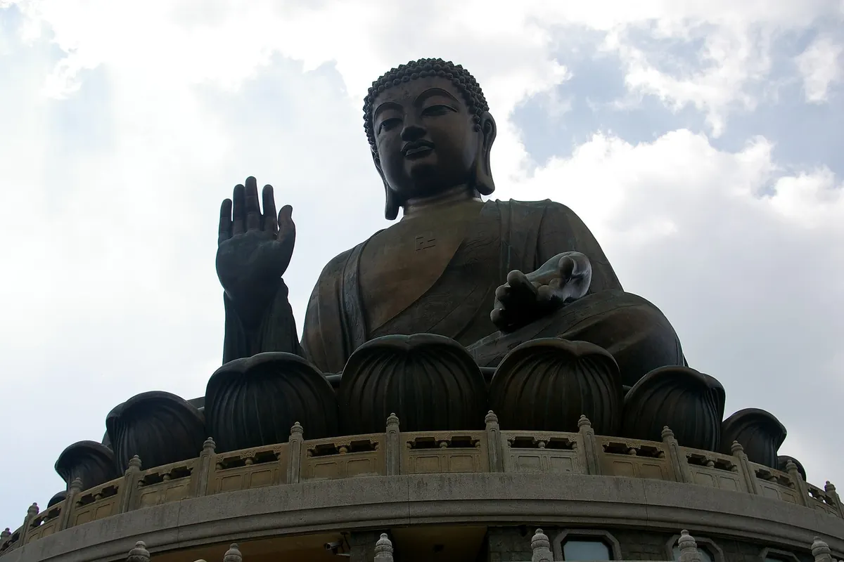 The bronze Tian Tan Buddha seen from below, one hand raised, seated on a lotus throne against a cloudy sky