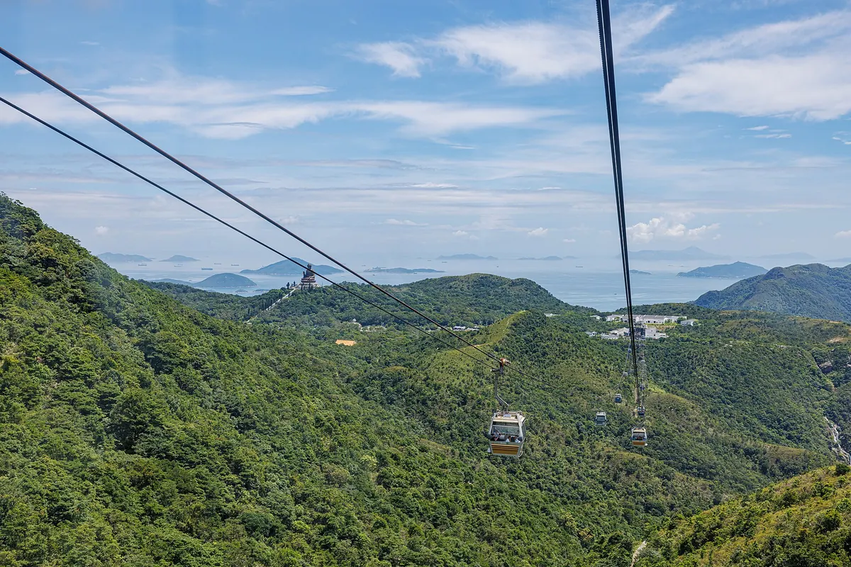 Two cables and a cabin crossing the wooded ridges of Lantau, with the Tian Tan Buddha on a distant hilltop and the sea and islands beyond