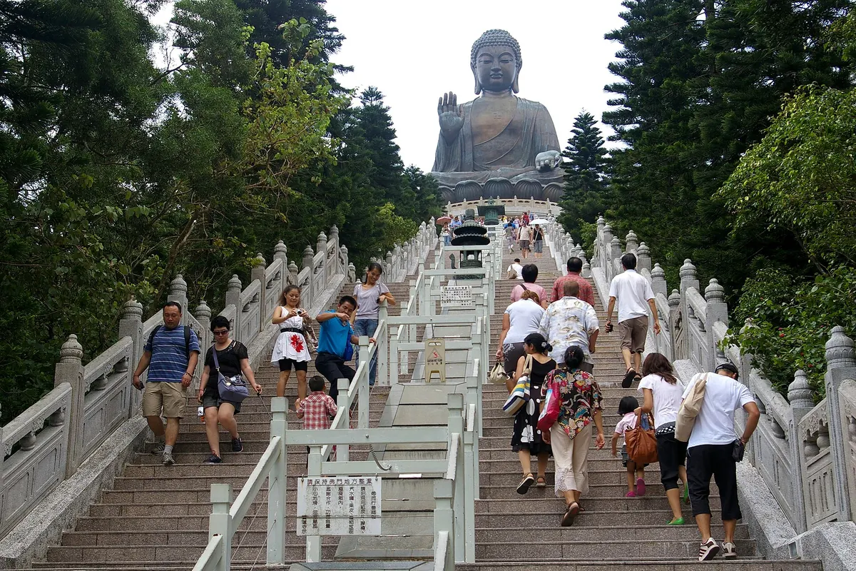 A wide stone staircase with white balustrades climbing towards the bronze Tian Tan Buddha, with visitors on the steps and trees on both sides