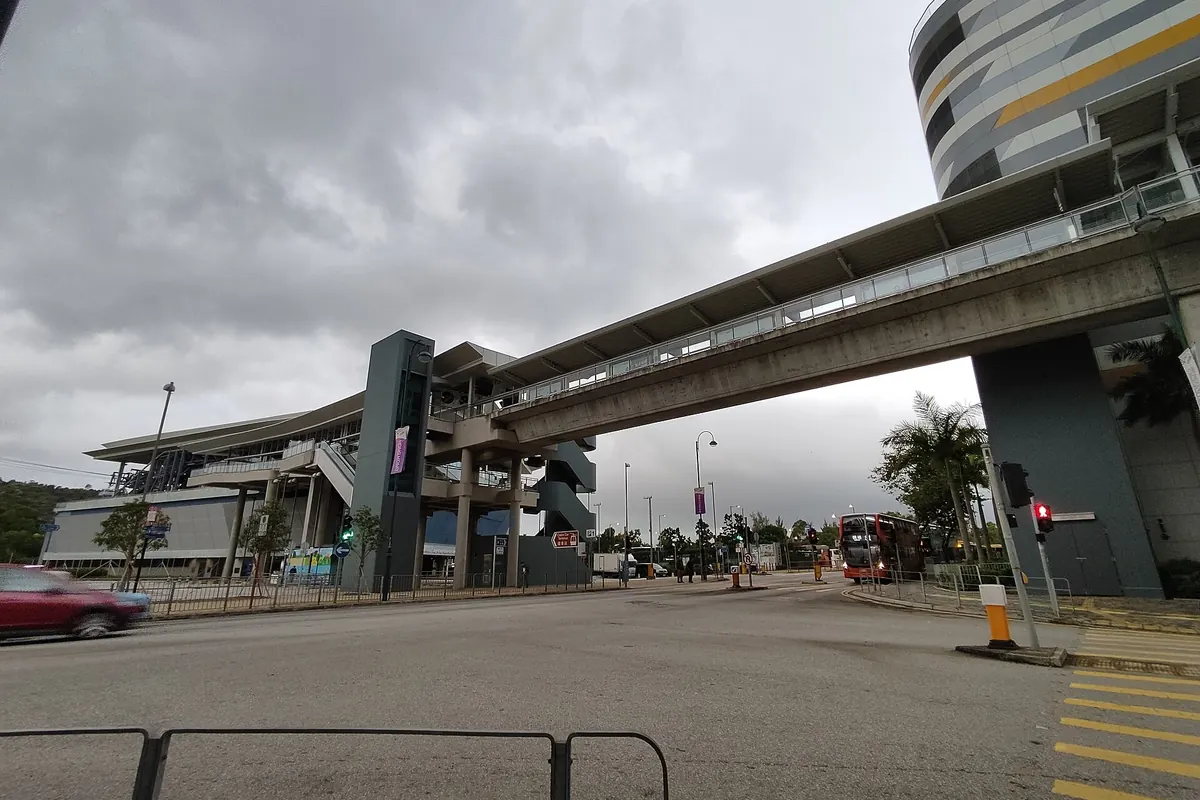 The low curved roof of the Tung Chung cable car terminal beside a pedestrian footbridge and a wide road under a cloudy sky