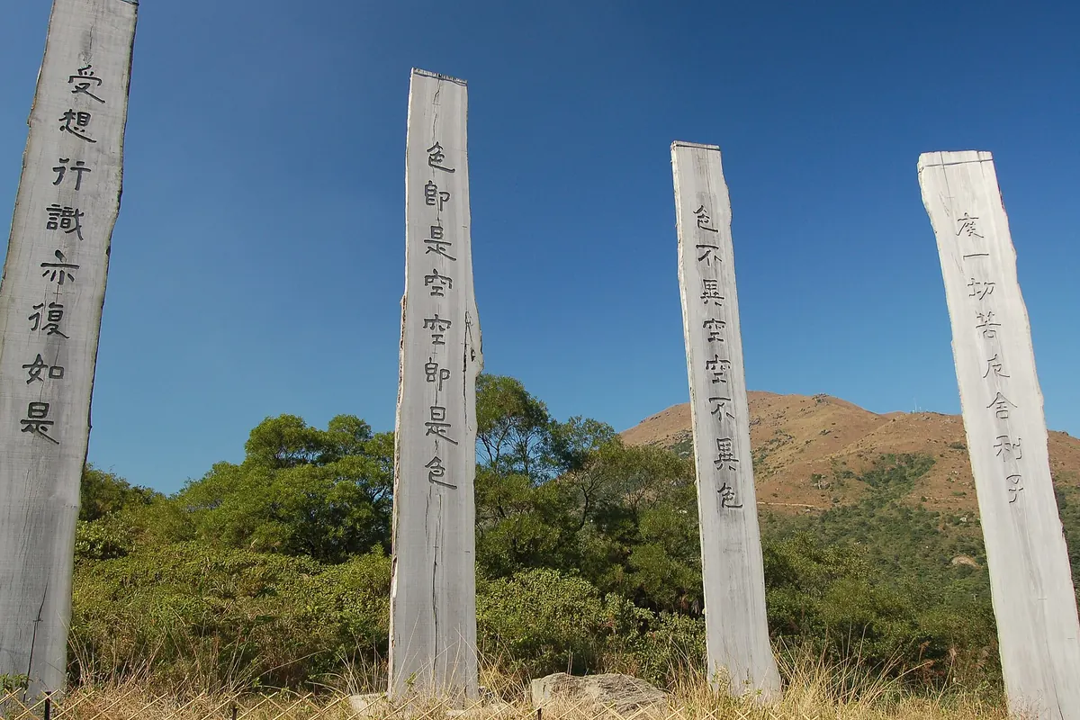 Four tall pale wooden columns carved with Chinese characters in a row against a deep blue sky, with a green hill behind
