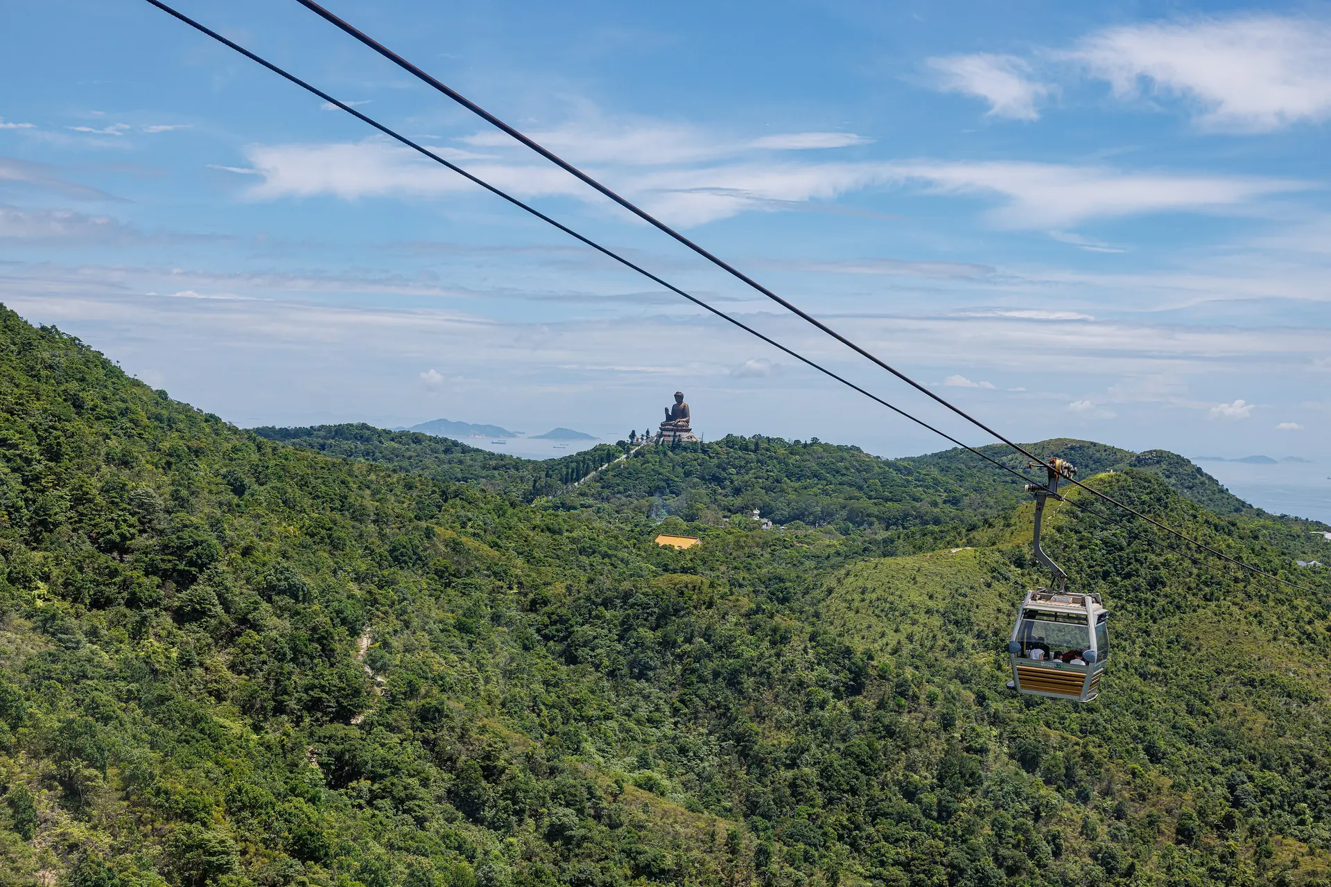 A cabin hangs from the cable over the wooded hills of Lantau, with the bronze Tian Tan Buddha on a distant hilltop and the sea and small islands beyond
