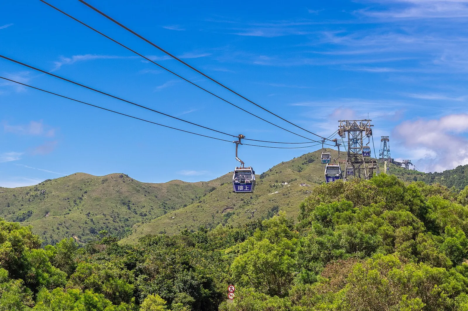 A blue cabin hangs from the cable above green hills, with a steel support tower and further cabins climbing away under a bright blue sky
