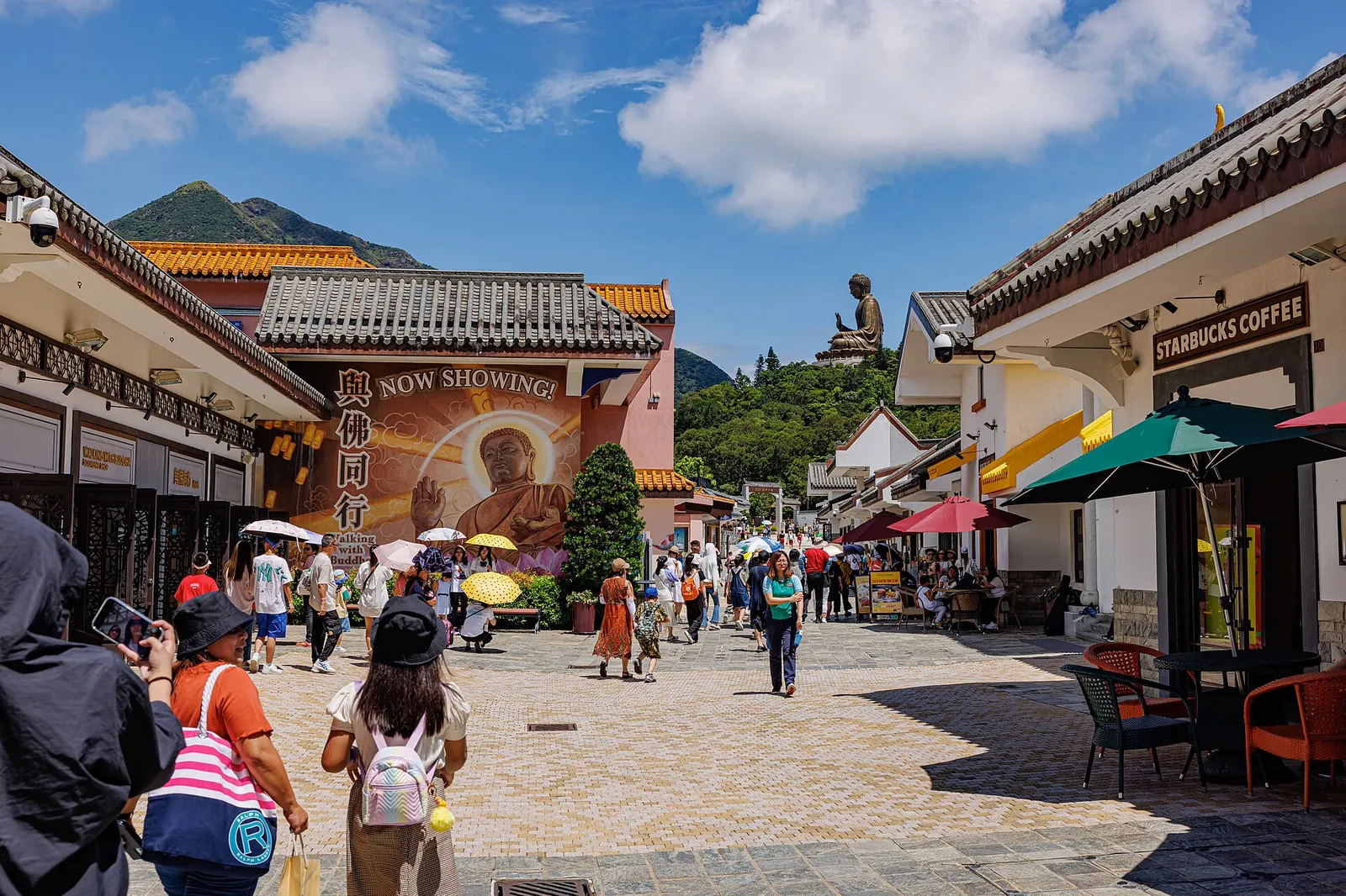 A paved street in Ngong Ping Village lined with low buildings with grey tiled roofs, red decorations and a hill rising behind